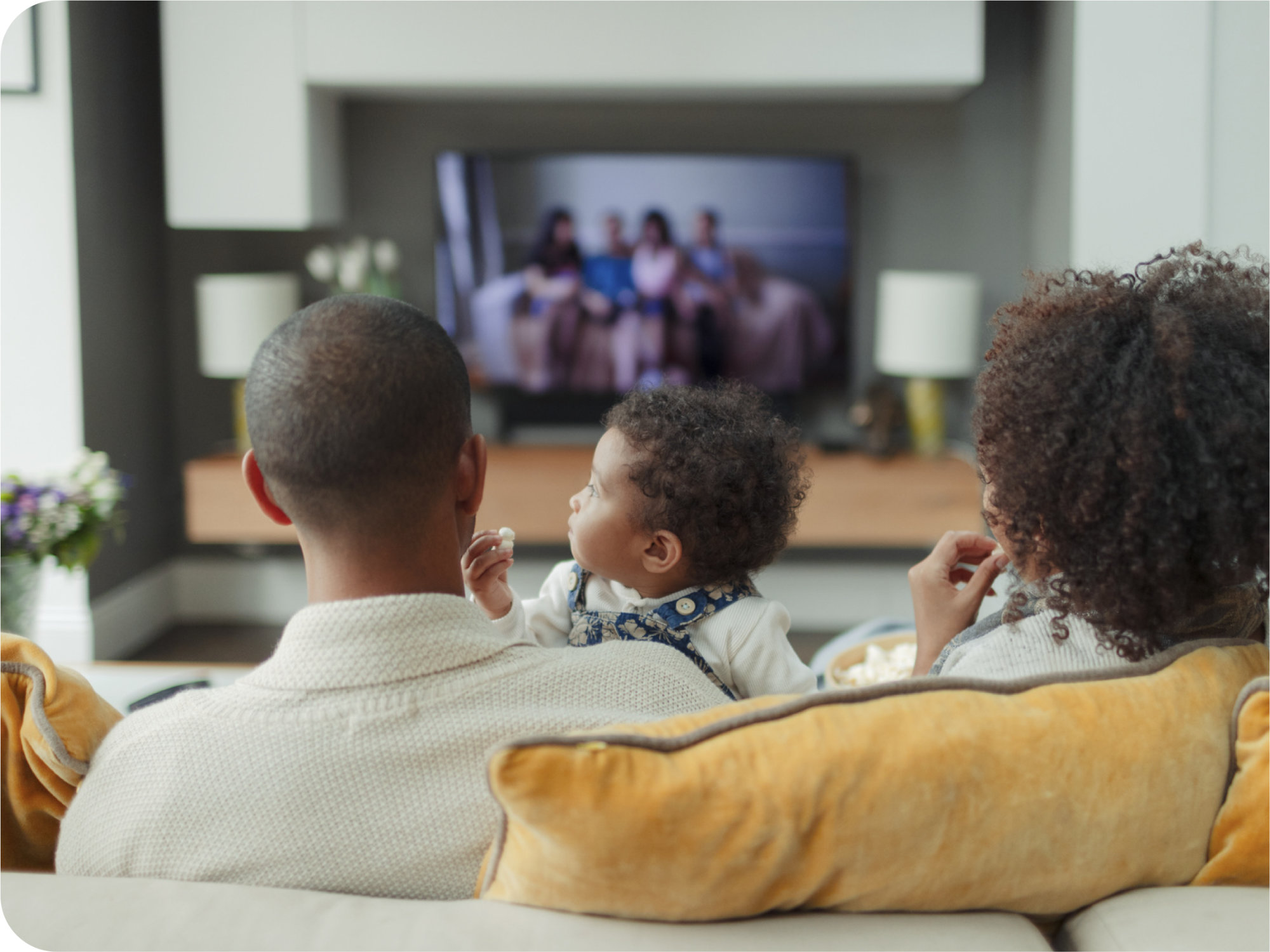 A man, woman and child, sit together on a yellow couch in their living room and watch television. The child, a toddler girl, sits on the man's lap and looks over her shoulder, towards the television screen. The woman and child eat popcorn.